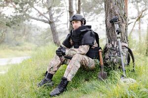 en man i en militär enhetlig och en skottsäker väst sitter i de skog nära en metall detektor och en militär ryggsäck. en man pausar i de arbete av minröjning de territorium foto