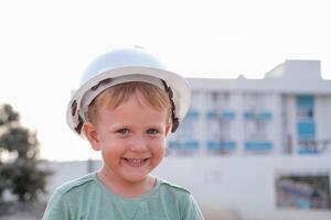en liten pojke i en vit hjälm utseende på de kamera och ler. en barn på de bakgrund av en konstruktion webbplats. yrke - byggare. foto