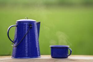 vattenkokare, blå emalj, och kaffe muggar på ett gammal trä- golv, suddig bakgrund av ris fält på soluppgång. foto