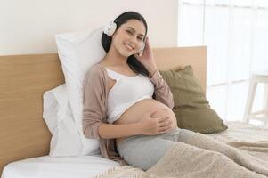 Lycklig gravid kvinna med hörlurar lyssnande till mozart musik och liggande på säng, graviditet begrepp foto