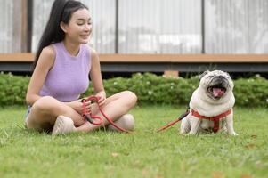 Lycklig asiatisk kvinna spelar med söt smart mops valp hund i de bakgård foto