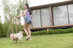 Lycklig asiatisk kvinna spelar med söt smart mops valp hund i de bakgård foto