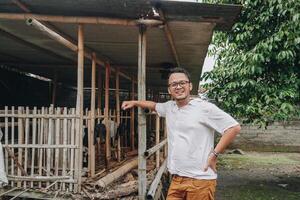 upphetsad ung asiatisk man stående i främre av traditionell bur tillverkad från trä och bambu i indonesien lantlig område med get inuti foto