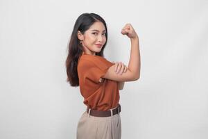 skön asiatisk kvinna bär en brun skjorta Framställ stark gest förbi lyft henne vapen och muskler medan leende stolt. foto