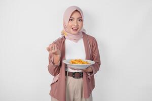 hungrig ung asiatisk muslim kvinna i hijab innehav gaffel och tallrik äter franska frites över isolerat vit bakgrund. foto