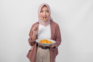 hungrig ung asiatisk muslim kvinna i hijab innehav gaffel och tallrik äter franska frites över isolerat vit bakgrund. foto