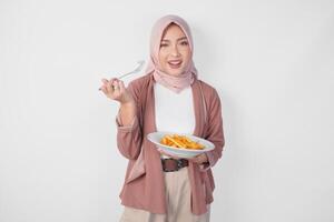 hungrig ung asiatisk muslim kvinna i hijab innehav gaffel och tallrik äter franska frites över isolerat vit bakgrund. foto