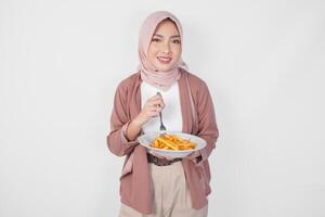 hungrig ung asiatisk muslim kvinna i hijab innehav gaffel och tallrik äter franska frites över isolerat vit bakgrund. foto