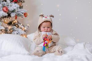söt bebis i en overall med rådjur horn spelar med en färgrik leksak Sammanträde nära de jul träd foto
