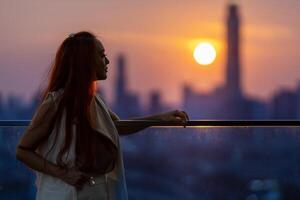 kvinna tittar och njuter av solnedgången från balkongen med solnedgången bakom skyskrapan i livlig stadskärna med ensamhet för ensamhet, ensamhet och drömmar om frihetslivsstil foto