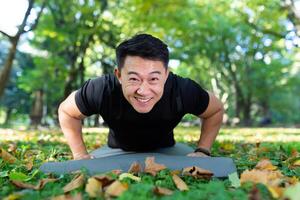 porträtt av ett idrottare i de parkera, ett asiatisk man utseende på de kamera och ler, en man i en sporter kostym gör fysisk övningar på en sporter matta. foto