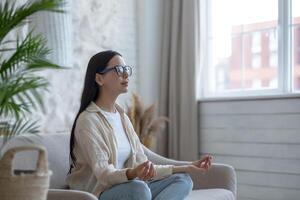 en skön brunett kvinna sitter på de soffa på Hem, vilar, mediterar i de lotus placera, gör yoga foto