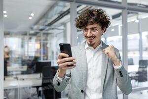 framgångsrik affärsman investerare använder sig av telefon, man fira seger och framgångsrik prestation, innehav hand upp triumf gest, chef i företag kostym inuti kontor stående nära fönster. foto