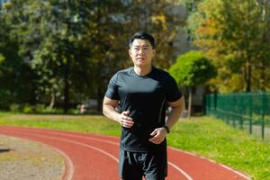 porträtt av en ung asiatisk man, en sportsman, ett idrottare löpning en maraton, håller på med en morgon- springa på en löpband i en stadion. foto