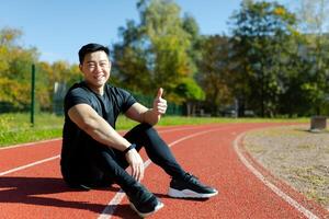 porträtt av en ung asiatisk manlig spotter, tränare, instruktör Sammanträde på en löpning Spår i en stadion, leende och pekande en super finger på de kamera. foto