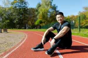 ung asiatisk sportsman, spotter, löpare Sammanträde i stadion på löpband och vilar efter joggning, träning. trött men leende, han utseende på de kamera. foto