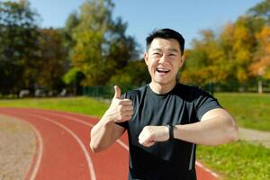 närbild porträtt av ett asiatisk ung man håller på med sporter och löpning i en stadion. han utseende på de smart Kolla på på hans hand, poäng en super finger, förtjust med de resultat och ler på de kamera. foto