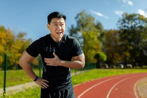 asiatisk man i sporter Träning har svår hjärta smärta, idrottare med hjärta ge sig på ge sig på i stadion innehav hand till bröst, på solig dag i kondition Träning och löpning. foto