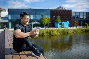 asiatisk sportsman vilar efter Träning Sammanträde på en bänk i de parkera nära de sjö och använder sig av de telefon, de man är leende och Lycklig handla om hans dagligen Träning utomhus på en solig dag. foto