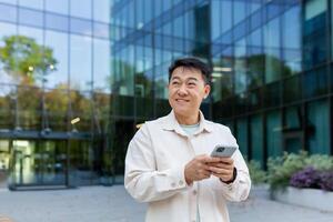 leende asiatisk man i tillfällig skjorta gående i stad använder sig av app på smartphone, man ser till sida på bakgrund av kontor byggnad från utanför. foto