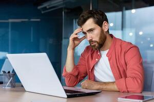 en betonade och trött ung affärsman, studerande, frilansare i en röd skjorta sitter i de kontor på en bärbar dator. innehav hans huvud. över tid arbete, deadline, problem. foto