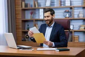 framgångsrik mogna man inuti kontor på arbete, senior man med skägg i Hem kontor mottagen post räv, Bank underrättelse, affärsman läsning Bra Nyheter i kuvert, nöjd med prestation resultat. foto