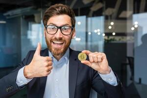 porträtt av mogna framgångsrik affärsman investerare, man visar kryptovaluta guld mynt till kamera och tummen upp fast, man Arbetar på stock utbyta inuti kontor uppkopplad användningar bärbar dator. foto