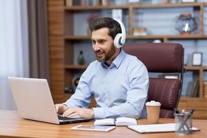 mogna affärsman med skägg och skjorta arbetssätt belåtet i kontor Sammanträde på skrivbord, man i hörlurar lyssnande till podcasts och ljudböcker på arbetsplats, chef skriver på bärbar dator. foto