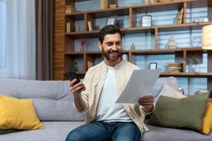 glad och leende man på Hem betalande räkningar och checkar, använder sig av bank app på telefon Sammanträde på soffa i levande rum innehav räkningar. foto