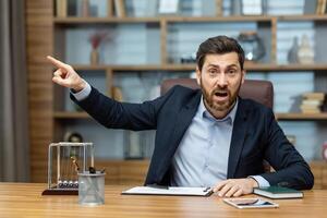 arg chef ropar på de kamera till de anställd poäng hans finger till de sida avsätter de underlydande, en affärsman i en företag kostym sitter på en tabell i en modern kontor. foto