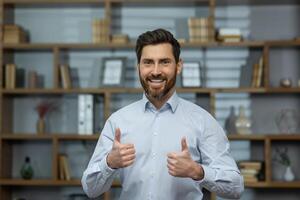 framgångsrik affärsman leende och ser på kamera, man visar tummen upp jakande och glatt håller med med prestation, chef i skjorta inuti kontor på arbete. foto