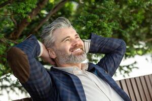 en 45 år gammal caucasian manlig i en mörk blå blazer och vit t-shirt, avkopplande utomhus med en belåten leende, utsöndrar en lugn och Lycklig humör. foto