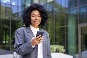 ung framgångsrik nöjd företag kvinna gående med telefon i händer, afrikansk amerikan kvinna i företag kostym med lockigt hår innehav smartphone, leende bläddring social media. foto