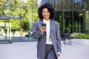 ung framgångsrik nöjd företag kvinna gående med telefon i händer, afrikansk amerikan kvinna i företag kostym med lockigt hår innehav smartphone, leende bläddring social media. foto