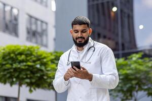 arab leende ung manlig läkare, studerande stående nära universitet sjukhus i enhetlig och använder sig av telefon. foto