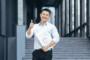 framgångsrik asiatisk affärsman utanför kontor leende, tummen upp jakande foto
