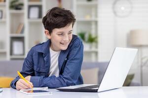 en ung kille, en studerande på Hem, sitter på en skrivbord med en bärbar dator, gör läxa och studier, ler belåtet, uppgifter uppkopplad kurser, avlägsen inlärning. foto