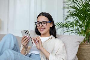 Lycklig ung studerande flicka i glasögon Sammanträde på soffa på Hem, innehav telefon, chattar och chattar med vänner, leende. foto