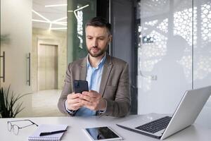 allvarlig koncentrerad mogna affärsman tänkande läsning uppkopplad social nätverk, erfaren mogna man innehav telefon använder sig av smartphone på arbetsplats inuti kontor med bärbar dator. foto