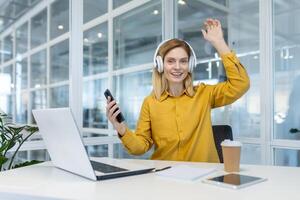 en glad kvinna professionell i en vibrerande gul skjorta vågor medan njuter musik på hörlurar, visa upp arbetsliv balans i en modern kontor miljö. foto