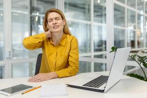kvinna i en gul skjorta känsla nacke smärta medan arbetssätt på en bärbar dator i en samtida kontor miljö. foto