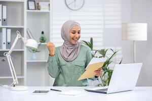 Lycklig kvinna i hijab fira medan läsning en brev på henne kontor skrivbord. hon är arbetssätt på en bärbar dator, som visar spänning och positiv känslor. foto