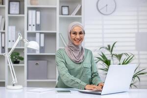 attraktiv muslim kvinna bär hijab och glasögon arbetssätt på trådlös bärbar dator på skrivbord i välorganiserad och ljus kontor miljö. klocka på vägg och grön växter i designer rum. foto