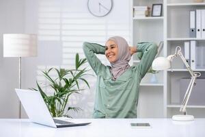 leende kvinna i hijab stretching på henne skrivbord i en modern kontor miljö med en bärbar dator, lampa, och grön växter. foto