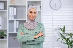 självsäker kvinna bär hijab med vapen korsade, stående i en modern kontor miljö. begrepp av bemyndigande, professionalism, och arbetsplats mångfald. foto