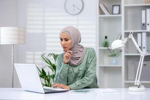 en tillägnad professionell bär en hijab är djupt fokuserade som hon Arbetar på henne bärbar dator på en ljus kontor skrivbord. foto