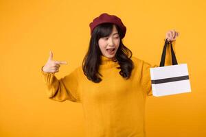 självsäker ung asiatisk kvinna i henne 30-talet, innehav en handla papper väska och pekande henne finger, visa upp henne mode stil i en gul Tröja och röd basker mot en gul bakgrund. foto