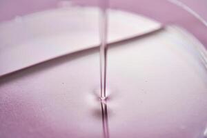 serum eller kosmetisk olja flöden in i en transparent skål på en lila bakgrund. foto