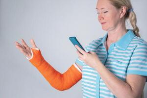 kvinna med en bruten rätt ärm i ett orange glasfiber kasta tittar på rehabilitering övningar på en smartphone, på en neutral bakgrund, hög kvalitet Foto