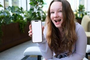 skön vit blond Tonårs modell flicka, mycket Söt, leende, naturlig tittar, utanför bär vit jacka som visar de smatphone på de kamera innehav den i hetero hand. mobil telefon i hand foto